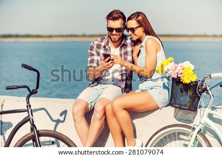 Look at this picture! Smiling young couple looking at mobile phone while sitting outdoors near their bicycles
