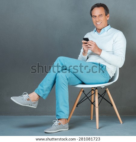Confidence and expertise. Happy mature man holding mobile phone and smiling at camera while sitting at the chair against grey background