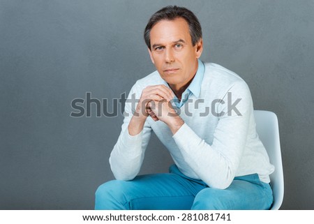 Serious and confident mature man. Thoughtful mature man keeping hands clasped and looking at camera while sitting against grey background