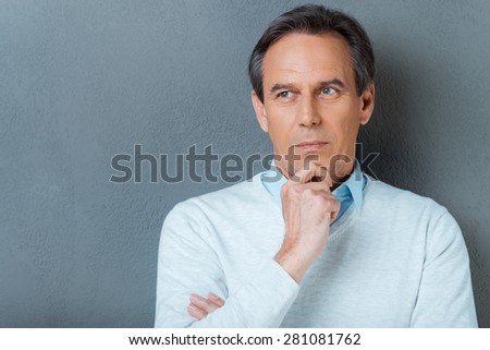 Lost in thoughts. Portrait of thoughtful mature man holding hand on chin and looking away while standing against grey background