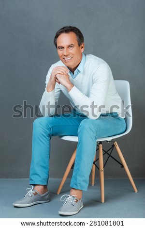He knows a secret of eternal youth. Happymature man keeping hands clasped and smiling at camera while sitting against grey background