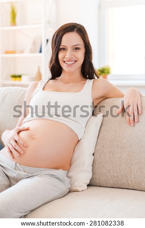 Looking forward to motherhood. Smiling pregnant young woman touching her belly and smiling at camera while sitting on sofa
