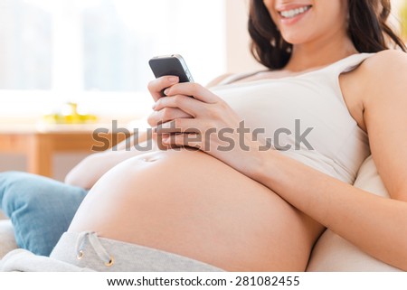 Keeping up to date.  Close-up of pregnant young woman holding mobile phone and smiling while sitting on sofa