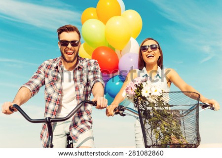 This is so much fun! Low angle view of cheerful young couple smiling and riding on bicycles with colorful balloons in the background