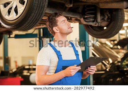Every detail is important. Confident young man in uniform holding clipboard and examining car while standing in workshop