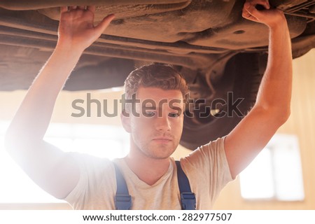 Confident mechanic. Confident young man looking at camera while standing underneath a car in workshop