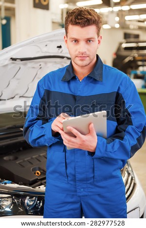 Concentrated on your problem. Confident young man working on digital tablet and looking at camera while standing in workshop with car in the background