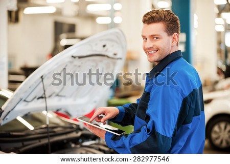 Years of mechanical experience. Confident young man working on digital tablet and smiling while standing in workshop with car in the background