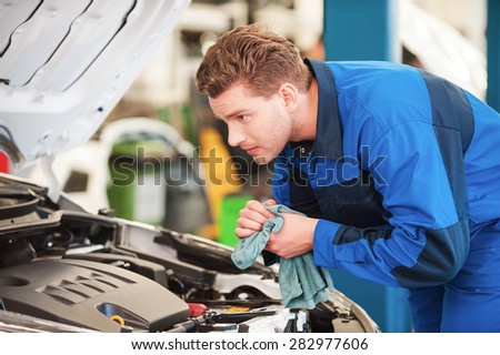 Mechanic at work. Concentrated young man in uniform examining car and wiping his hands with rag while standing in workshop