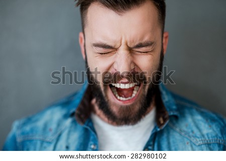 Unleashed emotions. Frustrated young man keeping eyes closed and mouth opened while standing against grey background