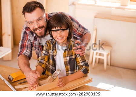 This is our happy place. Happy young male carpenter teaching his son towork with wood in his workshop