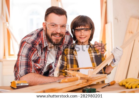 Carpentry is a family business. Cheerful young male carpenter embracing his son while leaning at the wooden table with diverse working tools laying on it