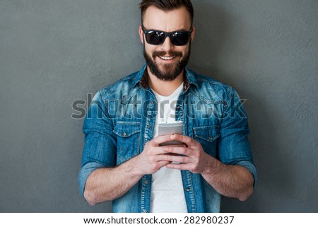 Texting to friend. Handsome young man holding mobile phone and smiling while standing against grey background