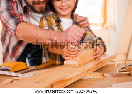 Sharing skills with his son. Close-up of smiling young male carpenter teaching his son to work with wood in his workshop