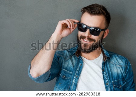 Rugged and manly. Smiling young man adjusting eyewear and looking away while standing against grey background