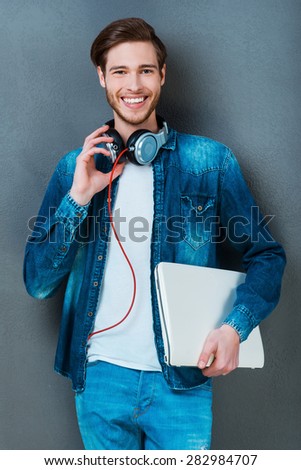 Everything I need is always in hand. Happy young man holding laptop and smiling at camera while standing against grey background
