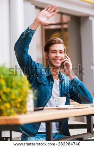 Meeting his friends in cafe. Smiling young man talking on the mobile phone and waving while sitting at sidewalk cafe