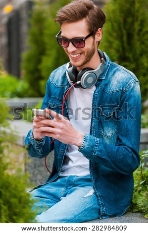 Spending carefree time outdoors. Handsome young man holding mobile phone and smiling while sitting outdoors