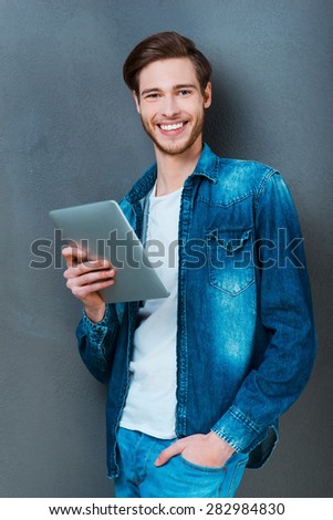 My digital friend. Happy young man holding digital tablet and smiling at camera while standing against grey background