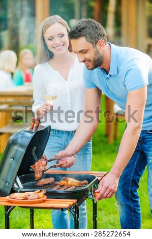 Need some help? Happy young couple barbecuing meat on the grill while other members of family sitting at the dining table in the background