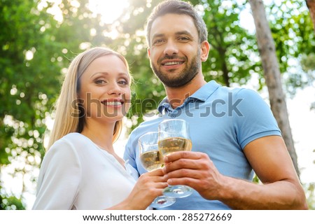 Happy loving couple. Low angle view of happy young loving couple holding glasses with white wine and smiling while standing close to each other outdoors