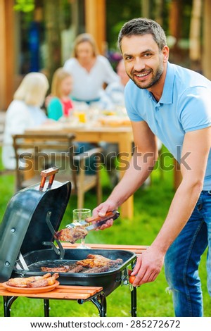 Enjoying family barbecue. Happy young man barbecuing meat on the grill and smiling while other members of family sitting at the dining table in the background