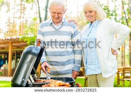 Enjoying barbeque together. Happy senior couple barbecuing meat on the grill while standing at the back yard of their house