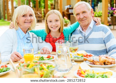 Enjoying time with grandparents. Happy little girl embracing her grandparents and looking at camera while sitting at the dining table outdoors together