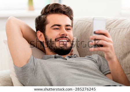 Happy to be at home. Smiling bearded man holding mobile phone and looking on it while laying on sofa