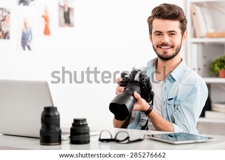 COnfident photographer. Cheerful young man holding digital camera and smiling while sitting at his working place