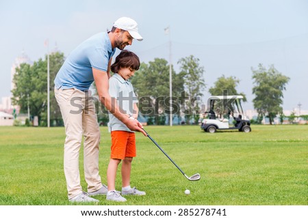 Sharing with golf experience. Cheerful young man teaching his son to play golf while standing on the golf course