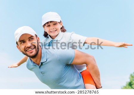 Having fun together. Cheerful young man carrying his son on shoulders while standing outdoors