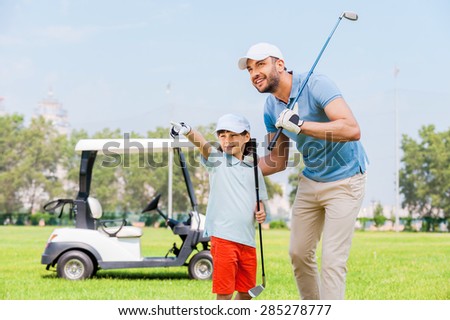 Look at that! Cheerful young man embracing his son and looking away while standing on the golf course