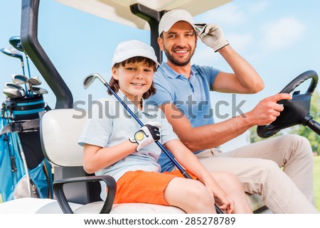 Two smiling golfers. Happy young man and his little son sitting in golf cart and looking at camera
