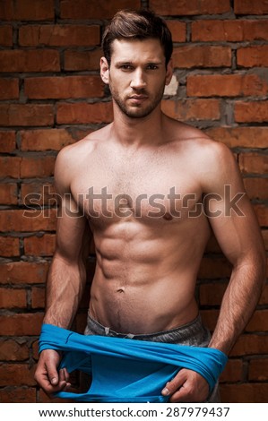 He is in a good shape. Serious young muscular man taking off his tank top while standing against brick wall