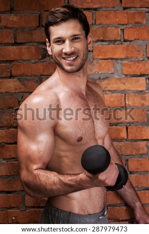 Muscular hadsome. Cheerful young muscular man training with dumbbells while standing against brick wall