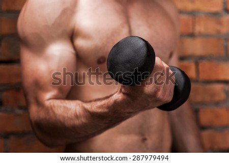 Strengthening his biceps. Close-up of young muscular man training with dumbbells while standing against brick wall