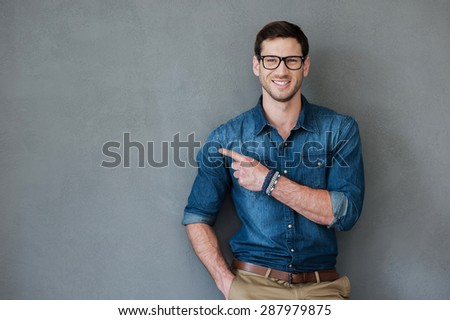 Pointing copy space. Joyful young man pointing away and looking at camera while standing against grey background