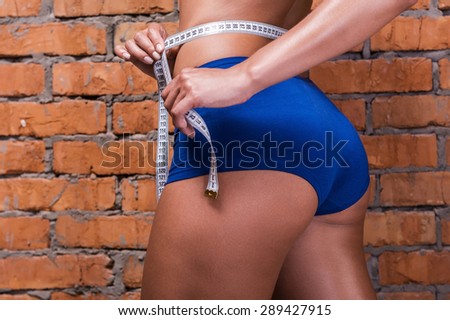 Keeping track on her weight loss. Close-up of beautiful sporty woman with perfect buttocks measuring her hip while standing against brick wall