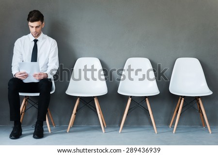 Waiting for interview. Confident young businessman holding paper while sitting on chair against grey background
