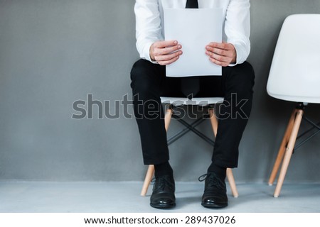 Waiting for interview. Close-up of young man holding paper while sitting on chair against grey background