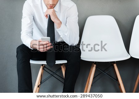 Staying in touch. Close-up of young man in shirt and tie holding mobile phone while sitting on chair against grey background