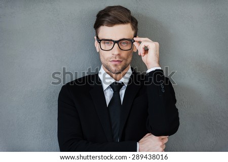 Stylish handsome. Confident young businessman adjusting his eyewear and looking at camera while standing against grey background