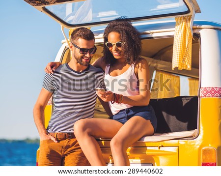 Look at this photo! Cheerful young couple looking at mobile phone together while both sitting at the trunk of retro minivan with sea in the background