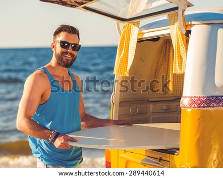 Happy to be at the seaside. Smiling young man taking skimboard out of car trunk and smiling at camera while standing on the beach