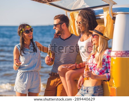 Enjoying summer day with friends. Group of cheerful young people discussing something and smiling while standing near their retro minivan with sea in the background
