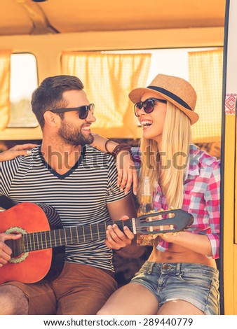 Spending carefree time together. Handsome young man sitting in minivan and playing guitar while his girlfriend bonding to him and smiling