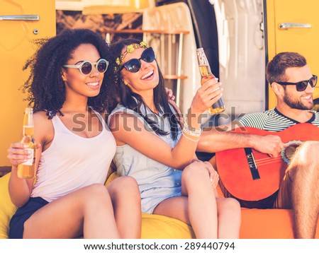 Carefree time with friends. Two cheerful young women holding bottles with beer and smiling while man playing the guitar with minivan in the background