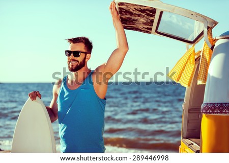 Ready to have some fun. Smiling young man holding skimboard and while opening a trunk door of his retro minivan with sea in the background