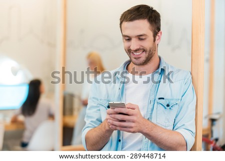 Staying connected. Happy young man looking at his mobile phone and smiling while his colleagues working in the background